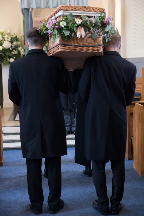 Pallbearers carrying floral-adorned wicker casket down aisle